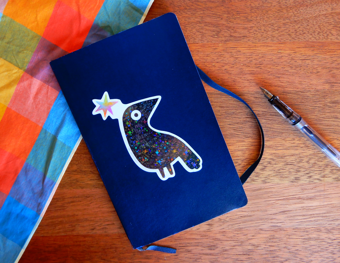 An image of an illustration of a black crow with holographic stars stuck on the front of a dark blue Moleskine notebook. It is resting on a wooden background, with a colourful checkered fabric to the left and an open fountain pen to the right.