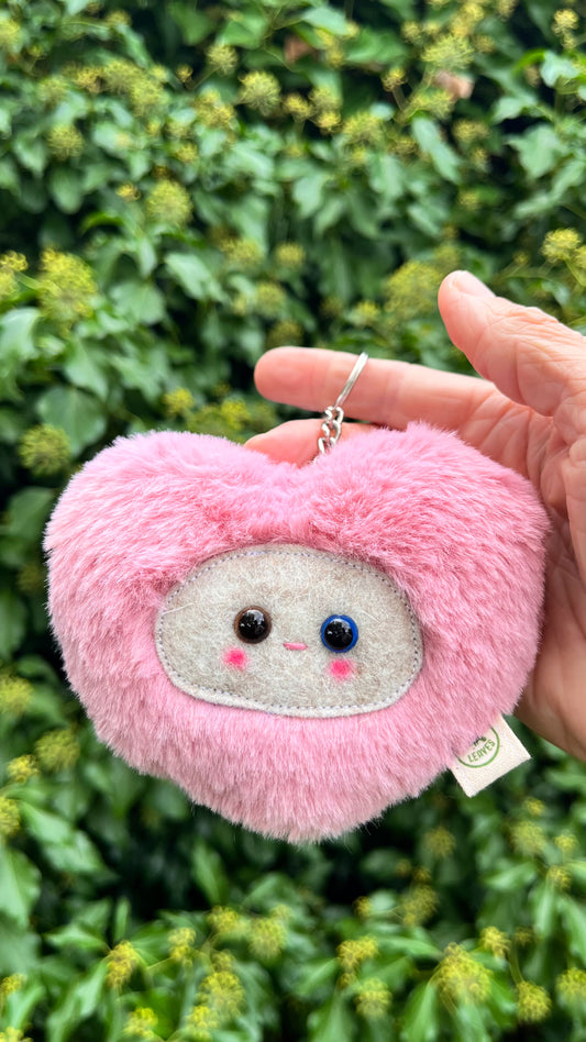 A pink fluffy heart shaped keychain/ bag charm with a light grey face and shiny eyes. 