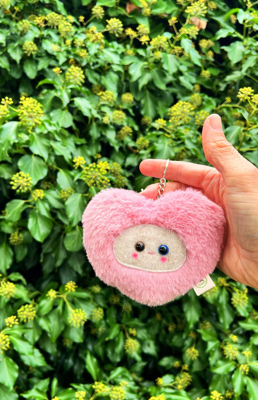 A pink fluffy heart shaped keychain/ bag charm with a light grey face and shiny eyes. 