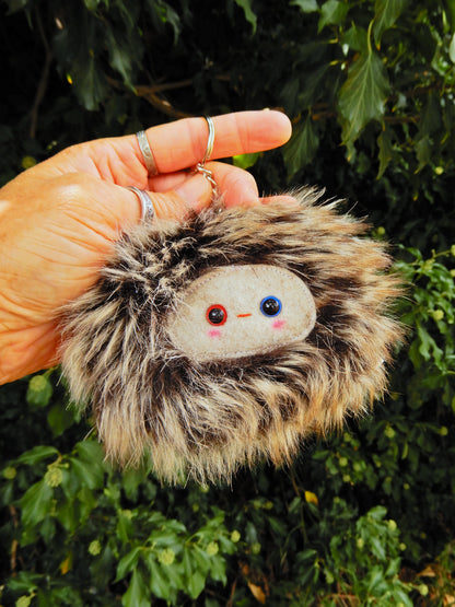 A brown fluffy plush key ring/ bag charm with a light grey face and pink cheeks.