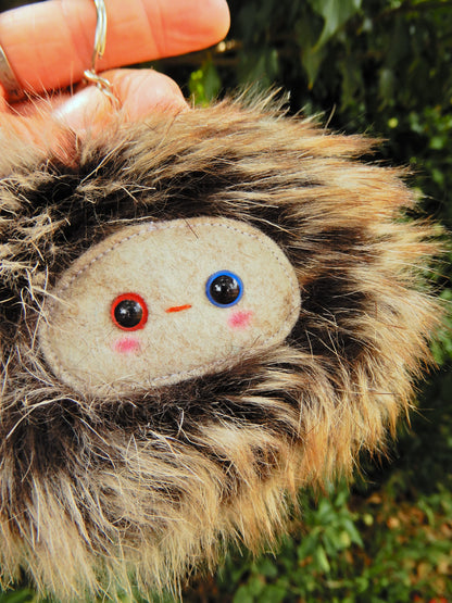 A brown fluffy plush key ring/ bag charm with a light grey face and pink cheeks.