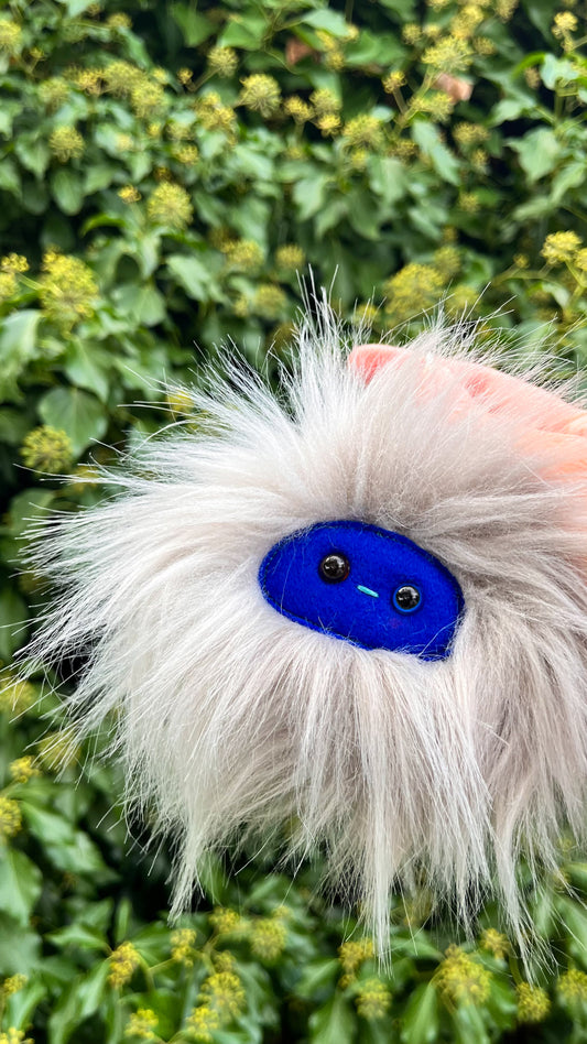A grey extra fluffy keychain/ bag charm with a  blue face and shiny eyes. 