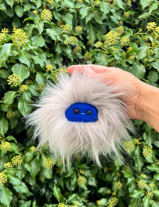 A grey extra fluffy keychain/ bag charm with a  blue face and shiny eyes. 