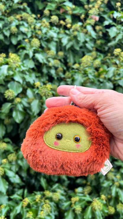 A rusty orange fluffy keychain/ bag charm with a green face and shiny eyes. 