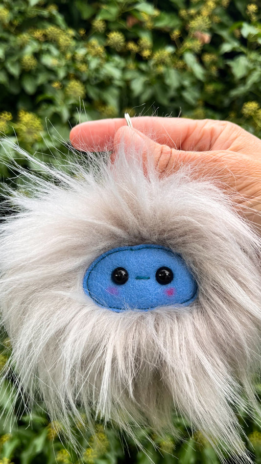 A grey extra fluffy keychain/ bag charm with a  blue face and shiny eyes. 