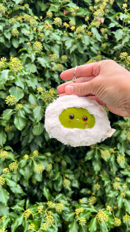 A white fluffy keychain/ bag charm with a green face and shiny eyes. 