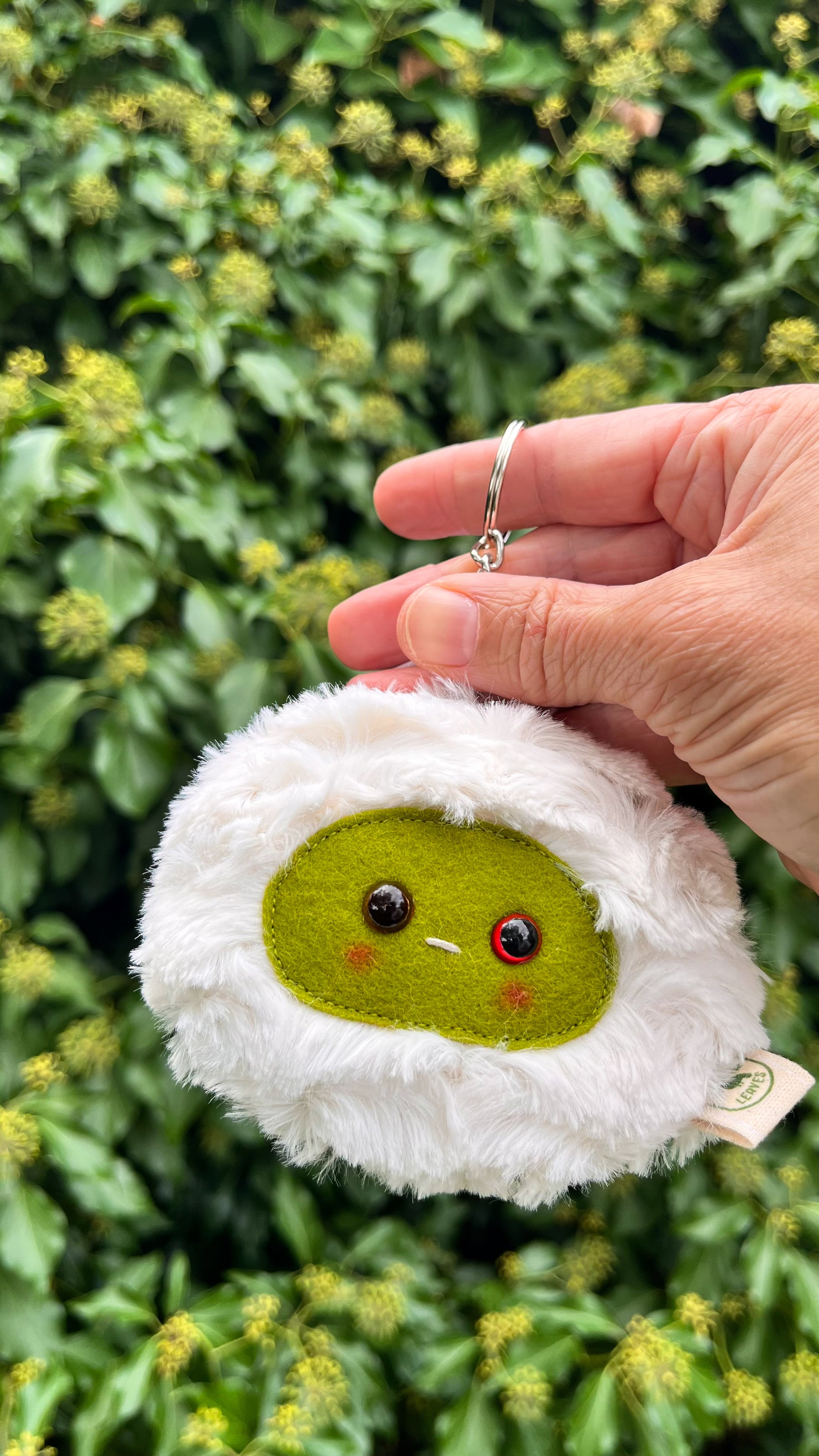 A white fluffy keychain/ bag charm with a green face and shiny eyes. 