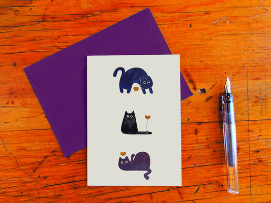 Greeting card with cat illustrations on a wooden surface with a purple envelope and pen.